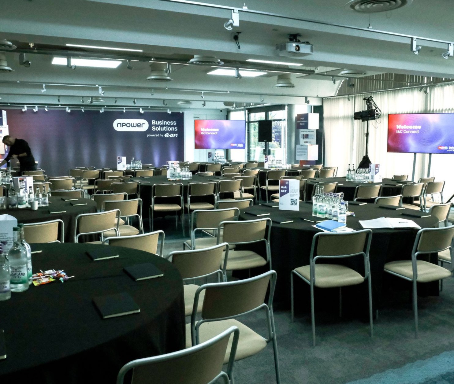 A conference room set up for an event, featuring rows of round tables with black tablecloths. Water bottles and notepads are on each table. A stage is in the background with large screens displaying a welcome message. The room is well-lit and has a modern design.