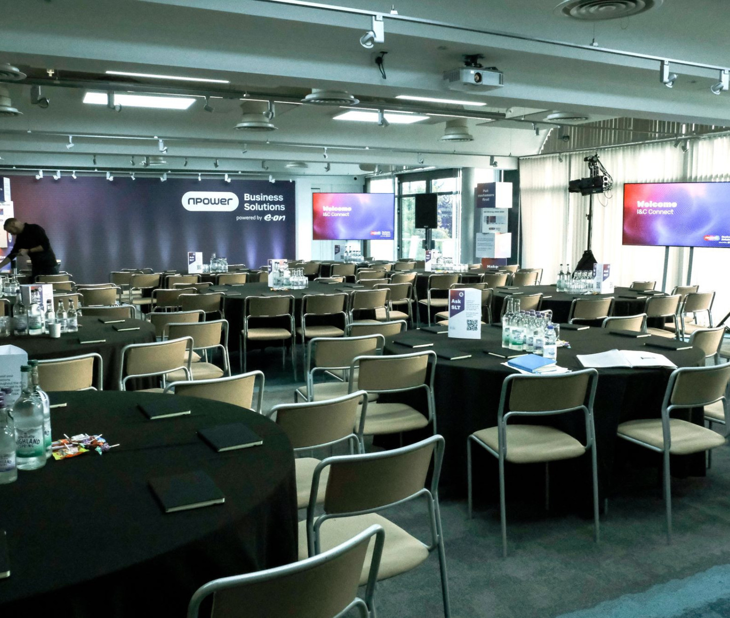 A conference room set up for an event, featuring rows of round tables with black tablecloths. Water bottles and notepads are on each table. A stage is in the background with large screens displaying a welcome message. The room is well-lit and has a modern design.