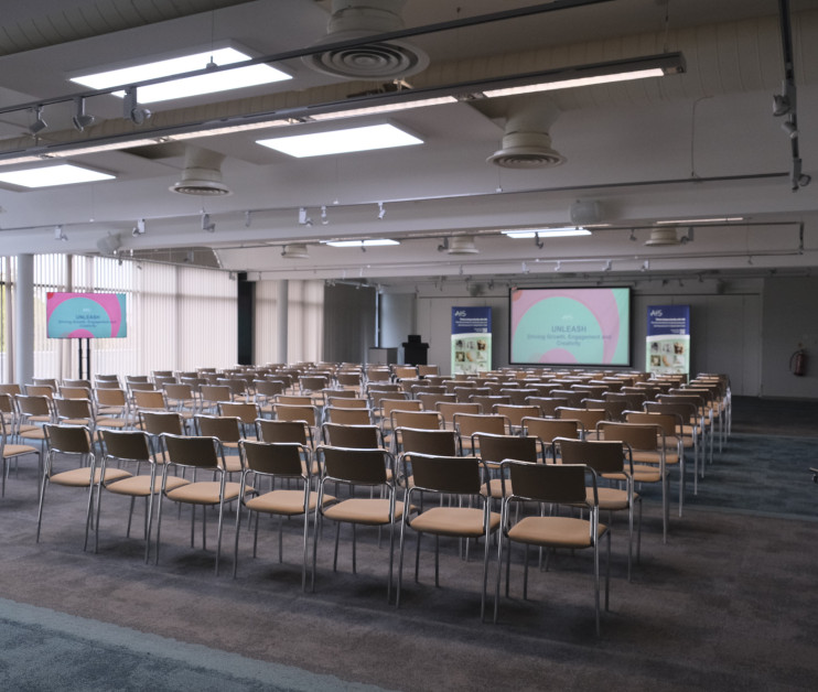 A large conference room with rows of beige chairs arranged in front of two screens displaying colorful presentations. The room has a modern design with large windows allowing natural light, and a blue carpet covering the floor.