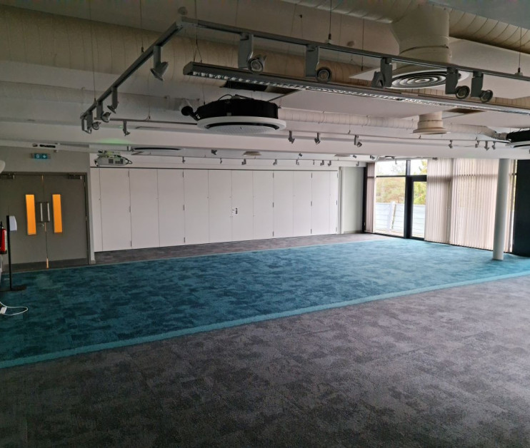 Empty meeting room with grey and teal carpeting, large windows allowing natural light, and storage cabinets along one wall. Ceiling features exposed ducts and lighting fixtures.