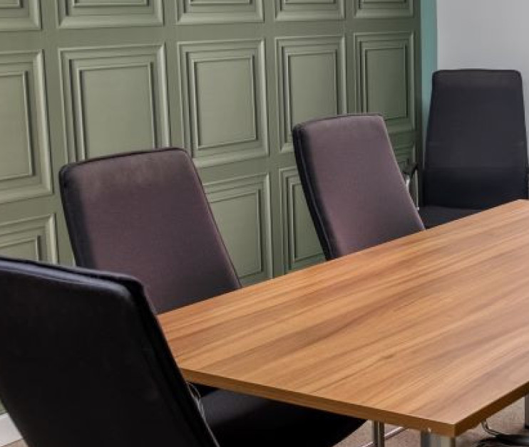 A modern conference room featuring a wooden table surrounded by six black chairs, with paneled green walls and a large window letting in natural light.