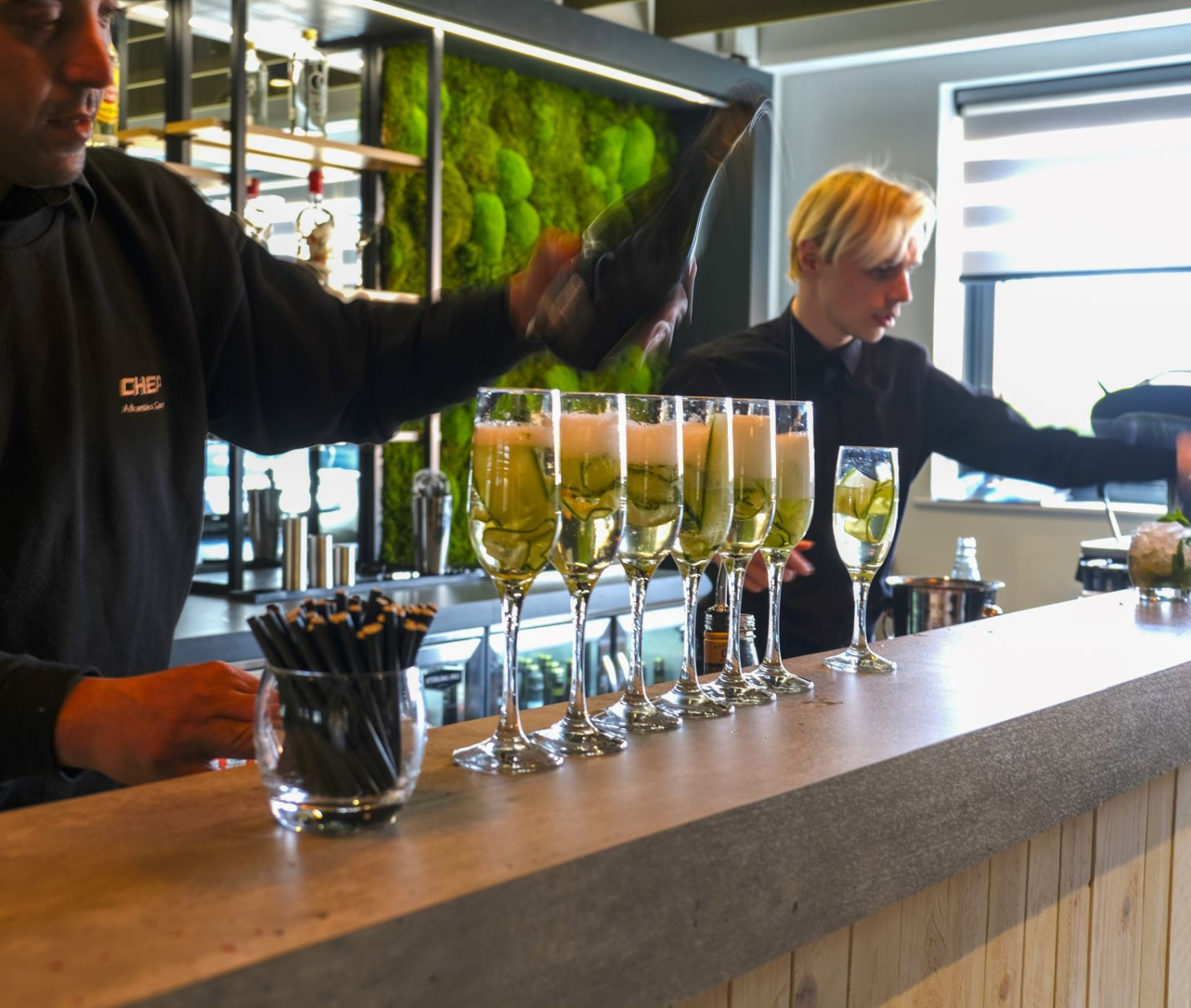 A bartender pours champagne into glasses lined up on a bar counter, while another staff member prepares drinks in the background. The bar features green moss decor, adding a natural touch to the modern setting.