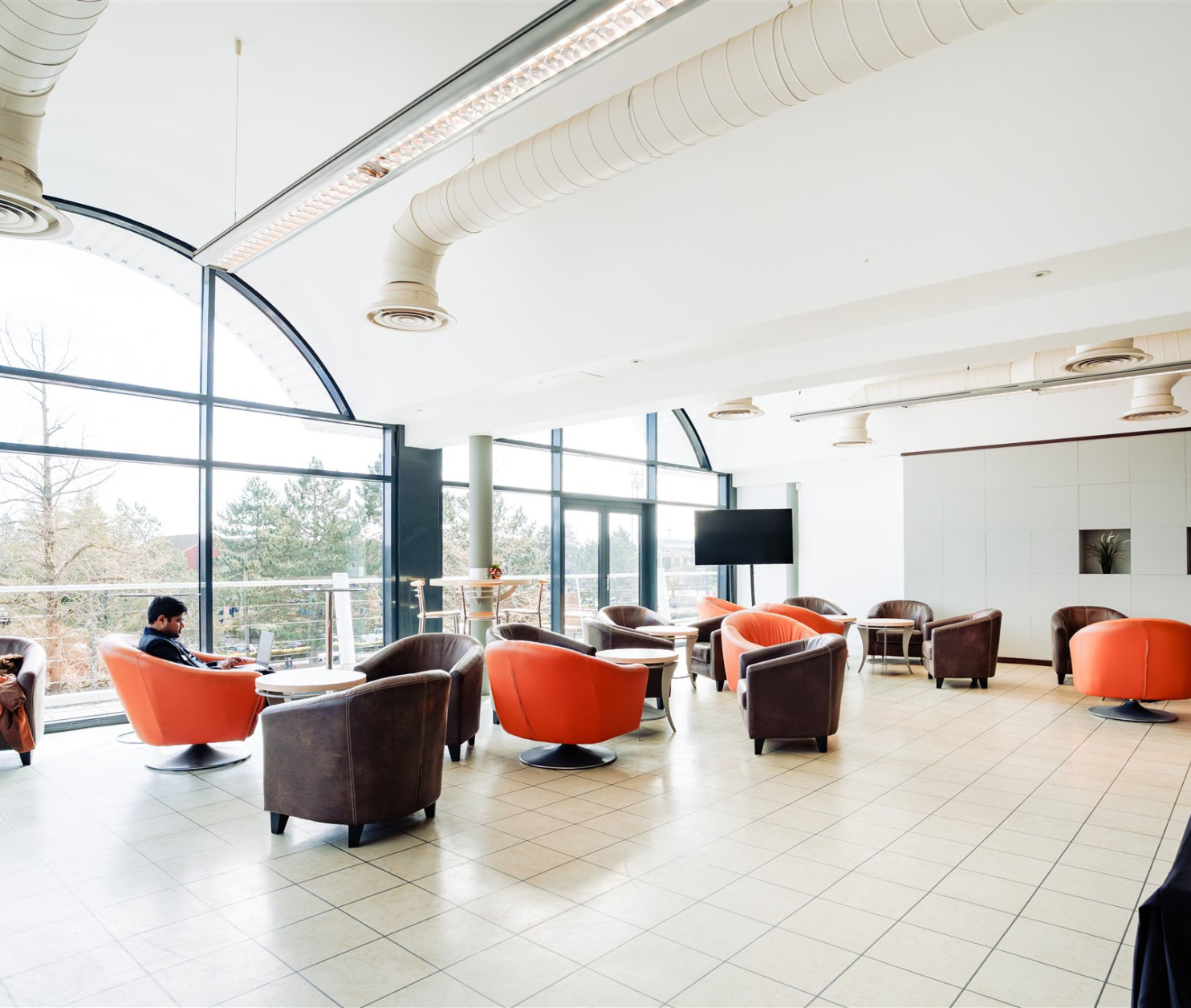 A spacious lounge area with large windows, featuring plush armchairs in orange and gray. A man is seated at a table, while another person is lounging on a chair nearby. The floor is tiled, and there is a modern design aesthetic.