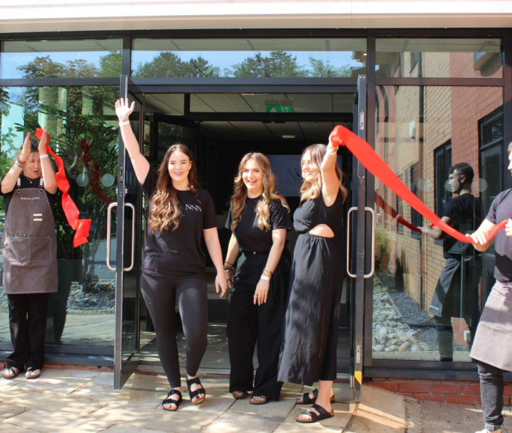 Two women smile and wave as they cut a red ribbon outside a modern building, celebrating a grand opening. In the background, two staff members applaud the event. All are dressed in black attire.