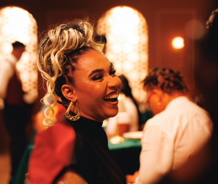 A woman with curly blonde hair and large hoop earrings smiles joyfully, sitting at a table in an elegantly decorated setting with warm lighting. Background figures are partially visible, engaged in conversation.
