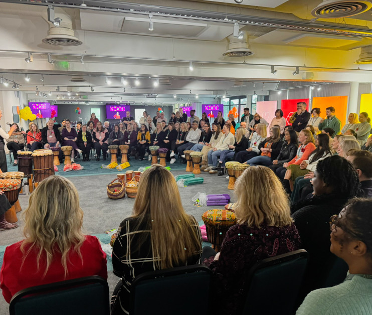 A group of diverse participants sits in a circle, engaging in a drumming session led by facilitators in a vibrant, well-lit room. Colorful decorations and screens are visible in the background, creating a lively atmosphere.