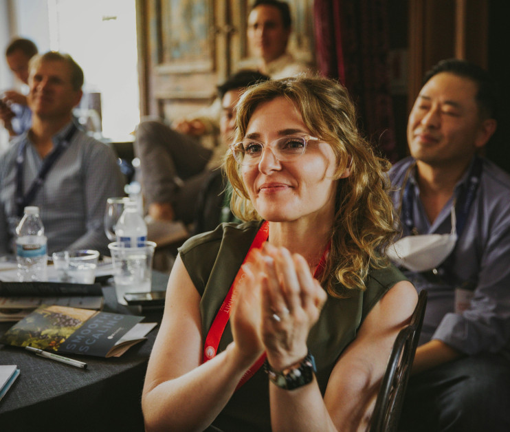 A woman with curly hair and glasses smiles while clapping, surrounded by an audience at a conference. Several people are seated at tables, with drinks and books visible in the foreground.