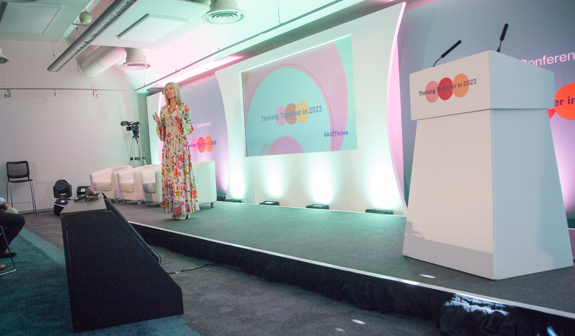 A speaker stands on stage at a conference, wearing a colorful floral dress. Behind her is a large screen displaying the event title 