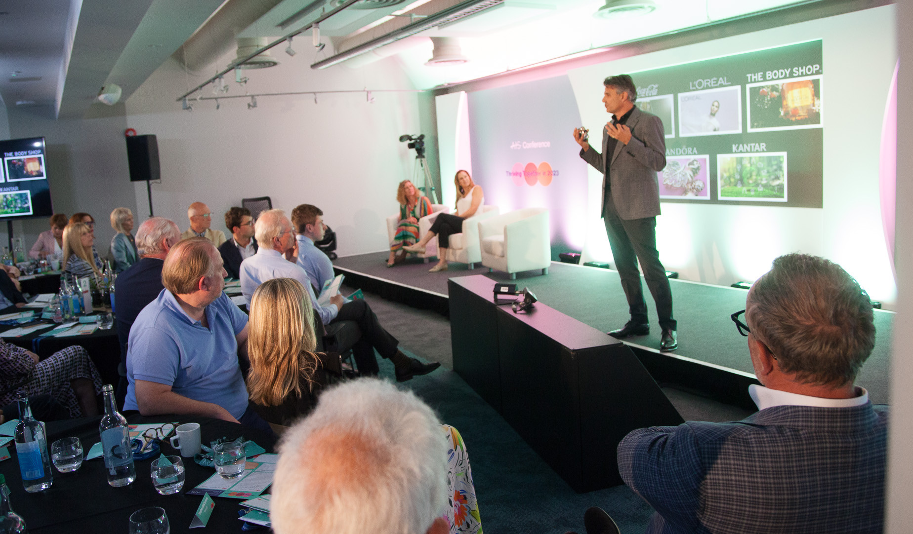 A speaker presents on stage at a conference, with a screen displaying logos of L’Oréal, The Body Shop, and Kantar. Attendees are seated at tables, engaged in the discussion.