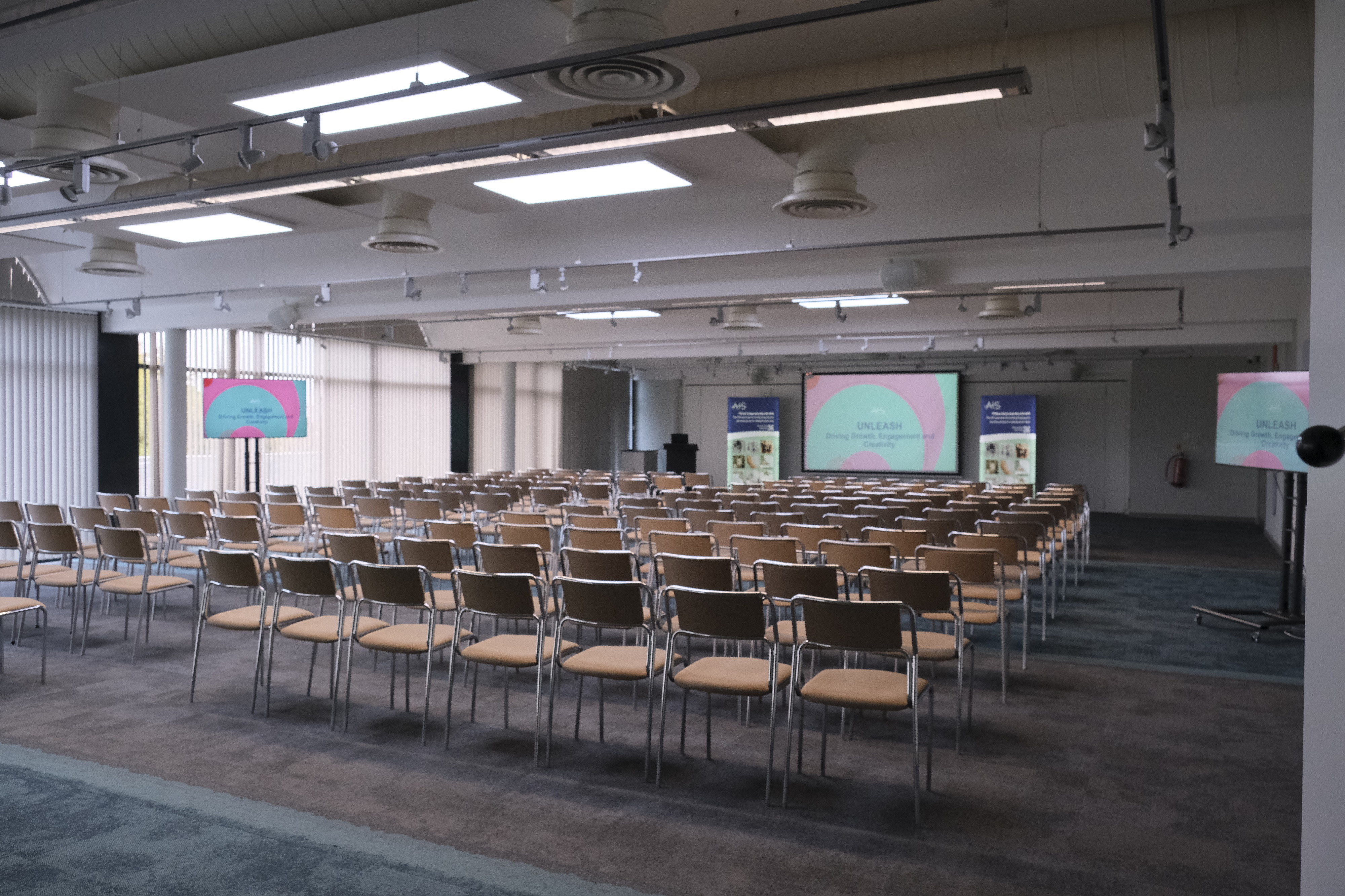 A spacious conference room set up with rows of chairs facing a stage. Two large screens display colorful presentations. Sunlight filters through large windows, illuminating the neutral-toned decor.
