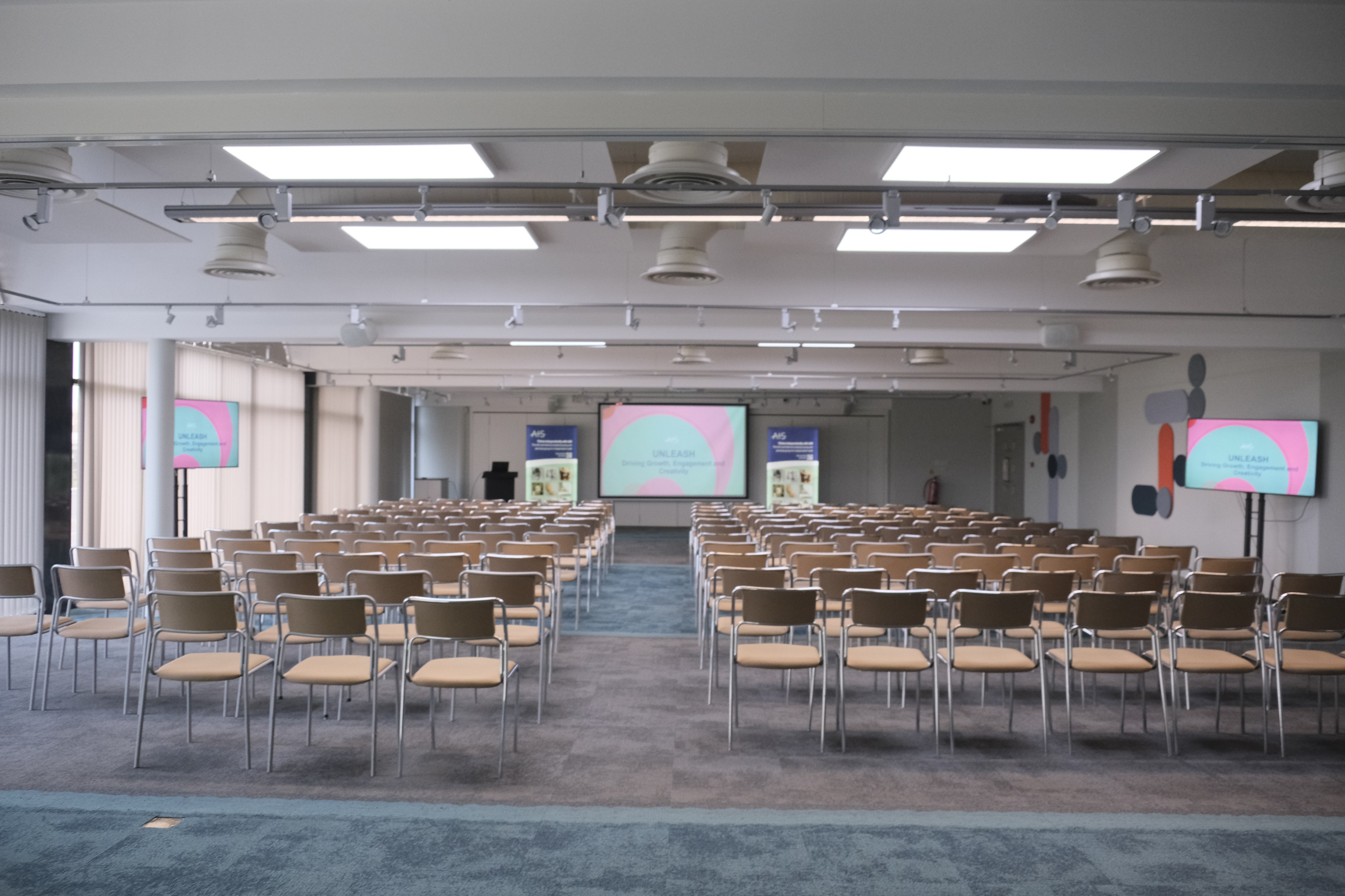 A spacious conference room set up with rows of beige chairs facing a stage. Two large screens at the front display pastel-colored presentation slides. Natural light fills the room through large windows, and the floor alternates between carpet and tiles.