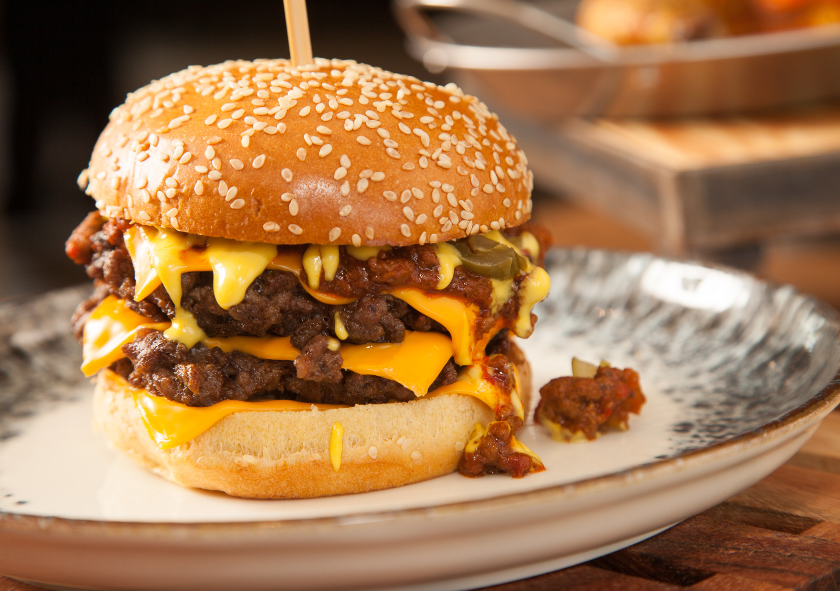 A towering cheeseburger stacked with two juicy patties, melted cheddar cheese, and topped with spicy jalapeños and a rich sauce, served on a sesame seed bun. A side of rustic fries is blurred in the background.