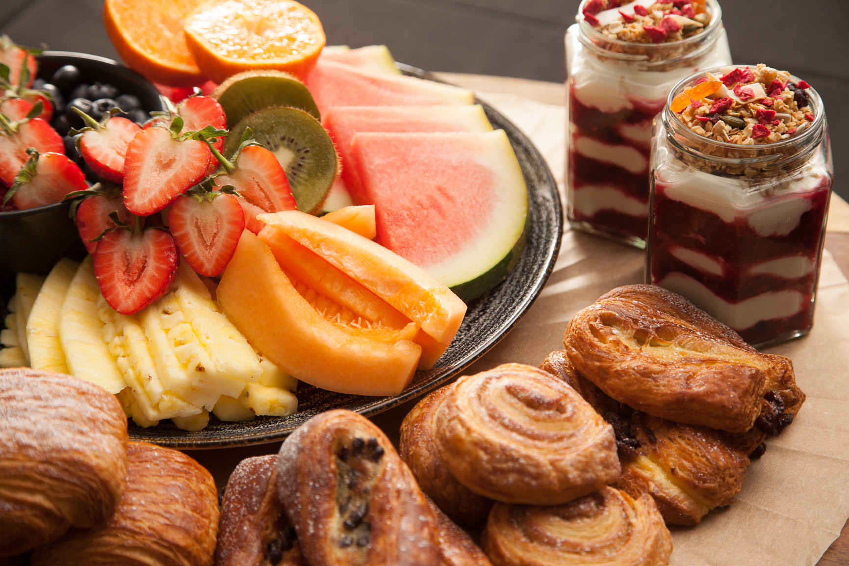 A colorful fruit platter featuring slices of watermelon, cantaloupe, honeydew, strawberries, kiwi, and oranges, alongside two jars of yogurt parfaits topped with granola and dried fruits. Fresh pastries, including croissants and cinnamon rolls, are also arranged nearby.