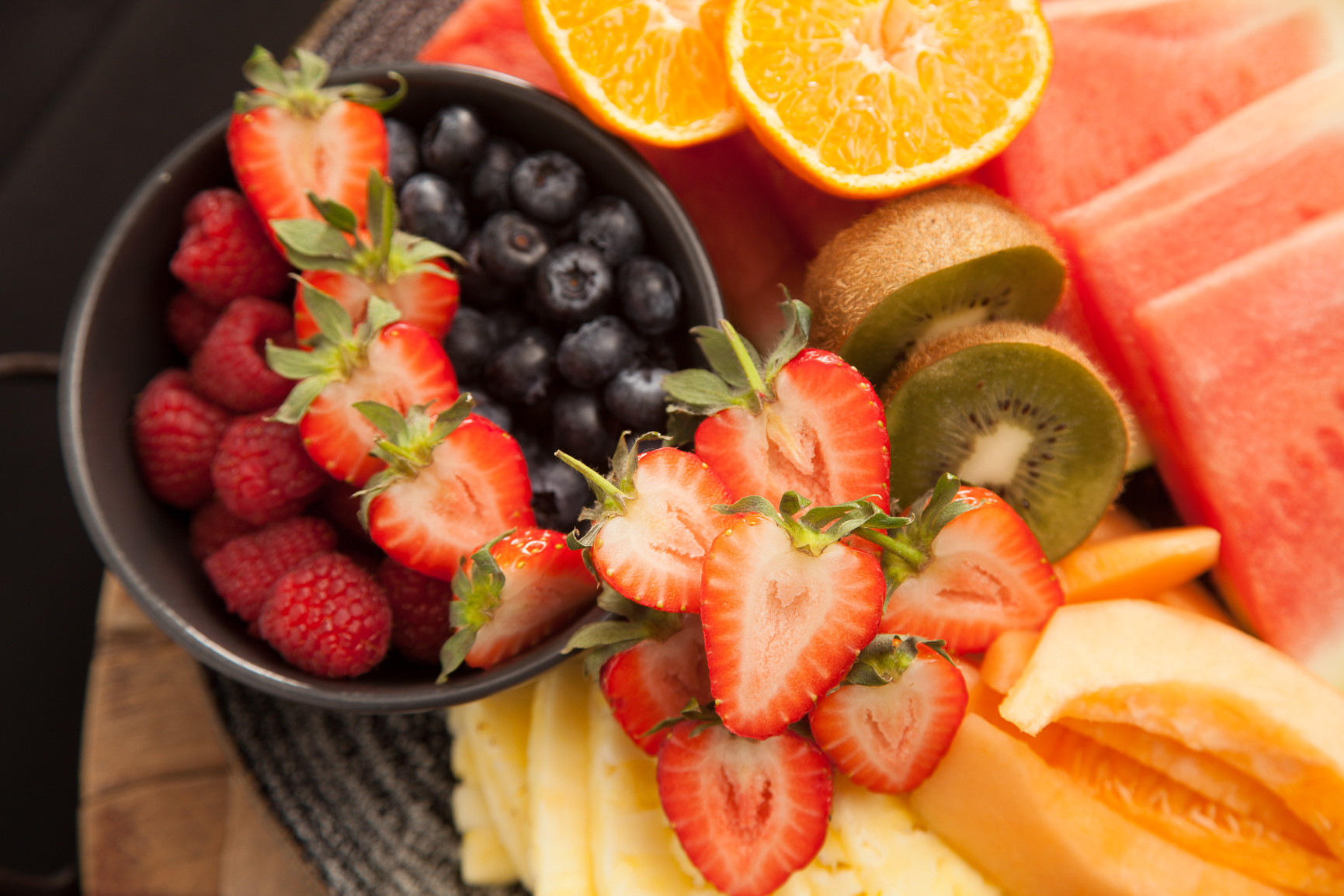 A vibrant fruit platter featuring a variety of fresh fruits. There's a bowl filled with blueberries and raspberries, surrounded by sliced strawberries, kiwis, watermelon, cantaloupe, pineapple, and orange segments.