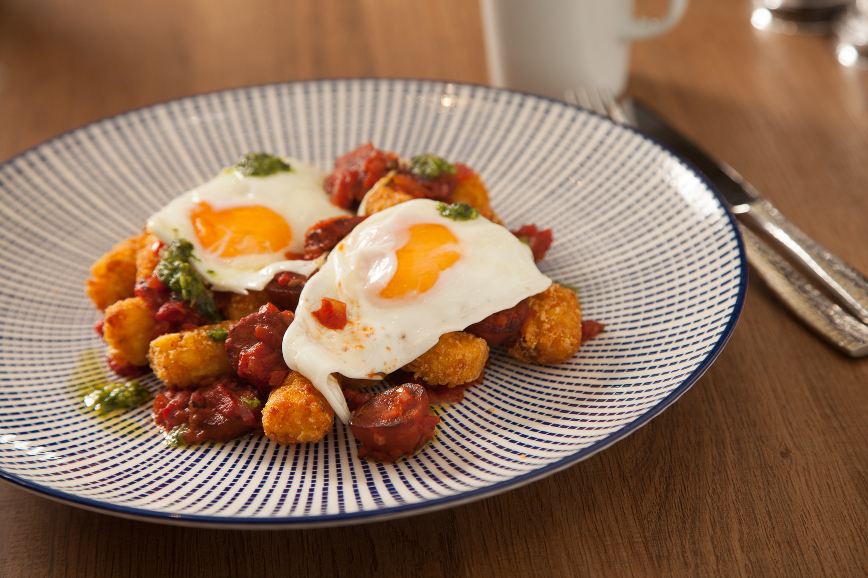 A plate of crispy tater tots topped with chorizo and two sunny-side-up eggs, garnished with a drizzle of green sauce and served on a decorative blue and white patterned plate.