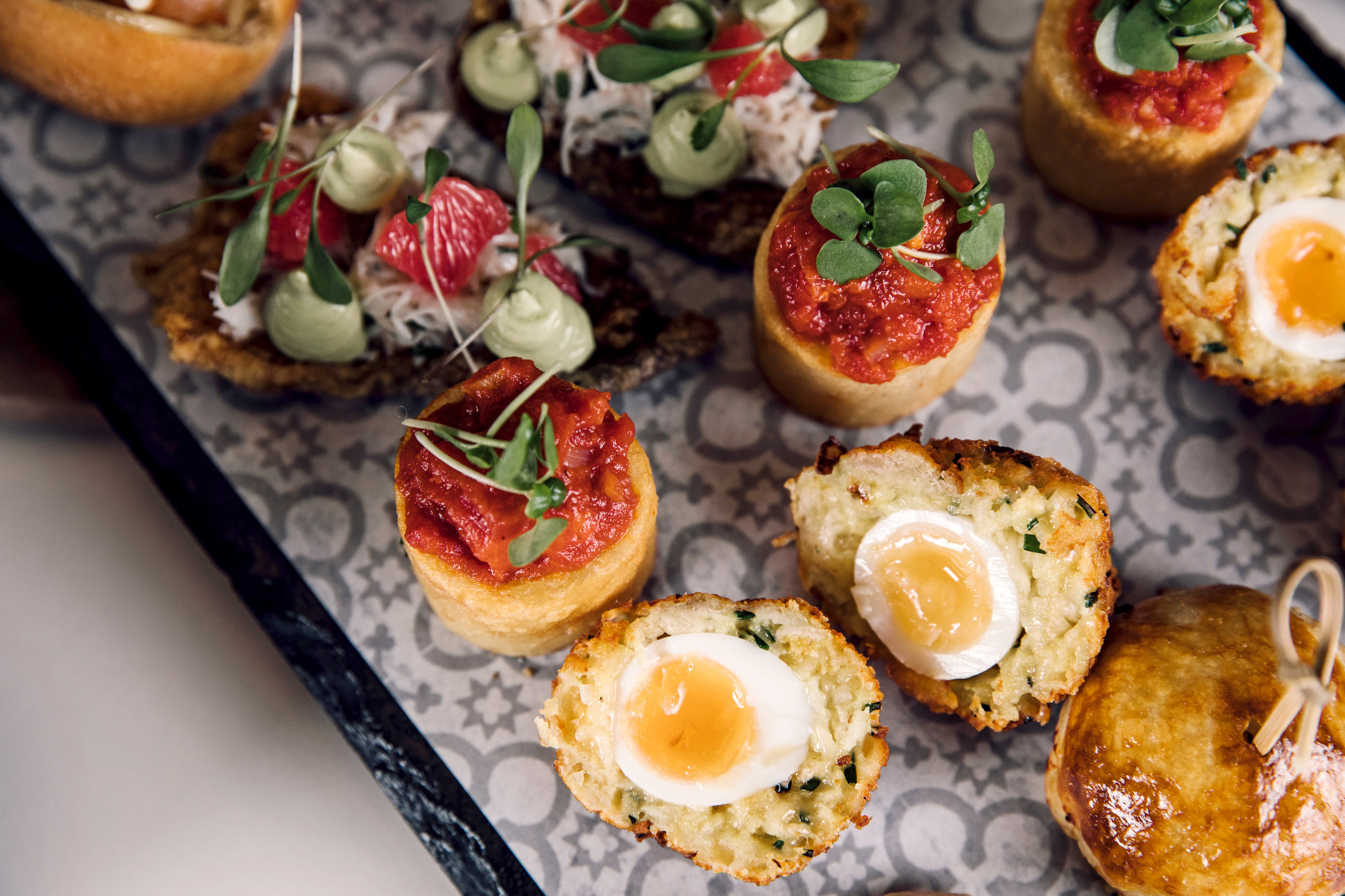 A platter of gourmet appetizers featuring various bite-sized treats. Some are topped with vibrant sauces and garnishes, while others have soft-boiled eggs nestled within crumbly bases. The arrangement is visually appealing with fresh herbs adding a pop of color.