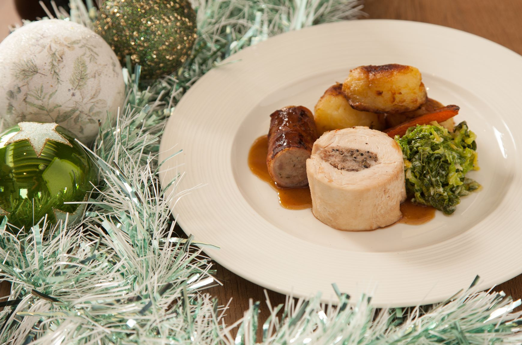 A festive plate featuring a slice of stuffed poultry, accompanying sausages, roasted potatoes, sautéed greens, and a drizzle of sauce. Decorated with sparkling green and silver ornaments and festive tinsel.