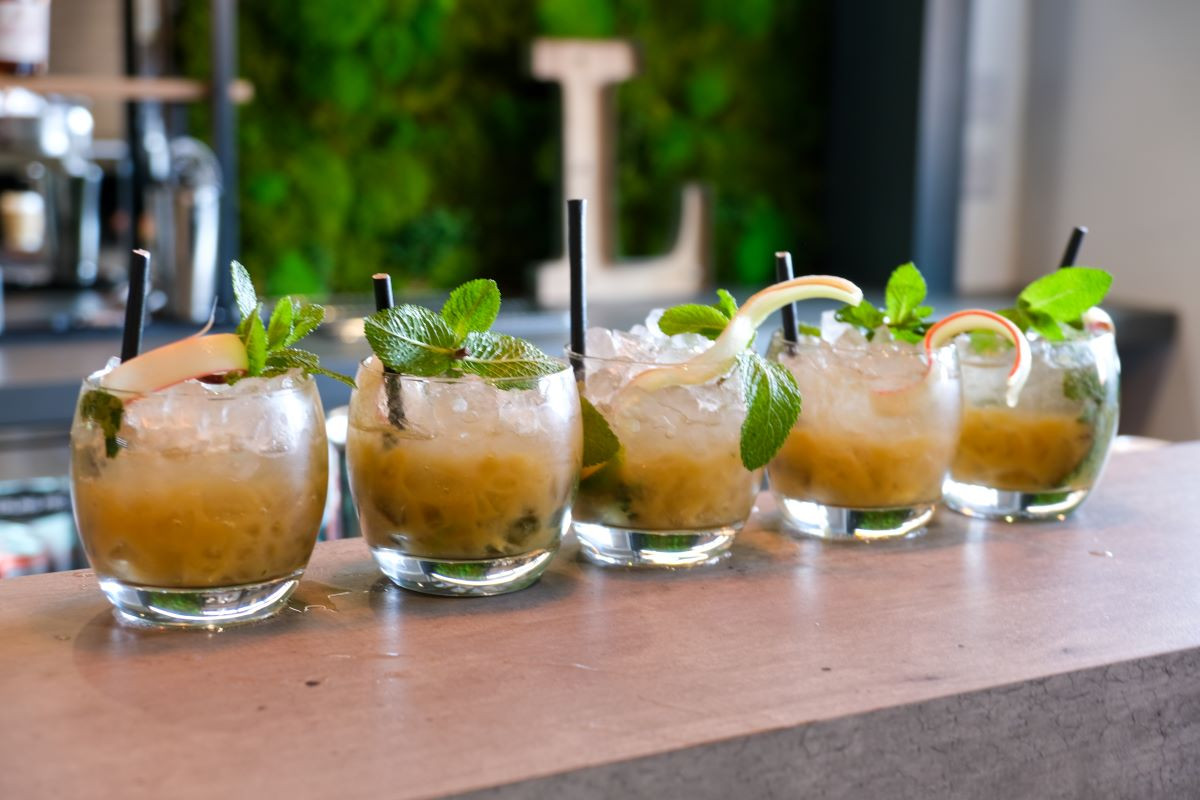 A row of six cocktails garnished with mint leaves and sliced fruit. Each drink is served in a clear glass filled with ice and a straw, set against a green moss backdrop.
