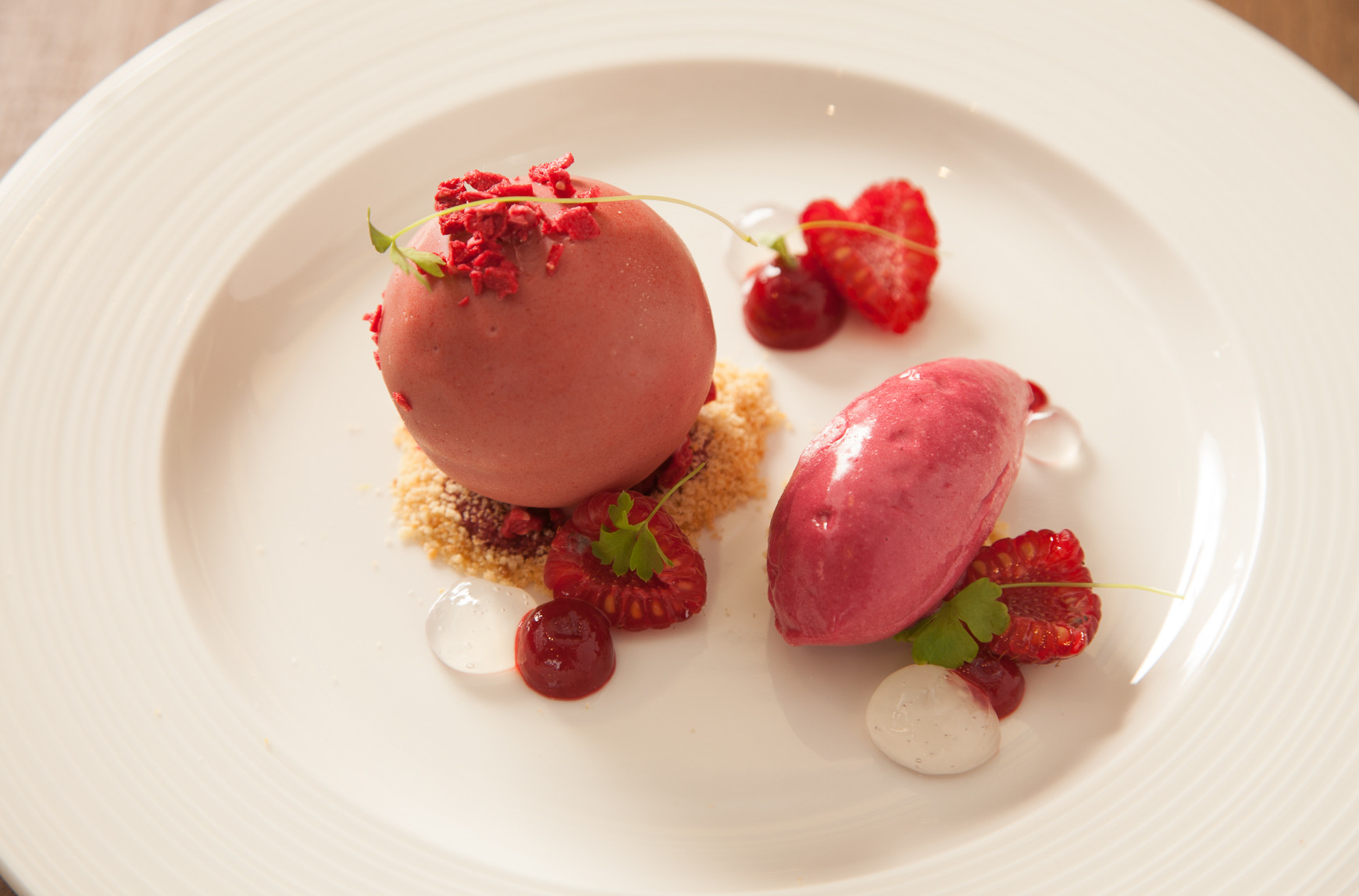 Two scoops of vibrant pink sorbet are elegantly plated with fresh raspberries, edible flowers, and dollops of cream. Crushed cookie crumbs add texture to the presentation.