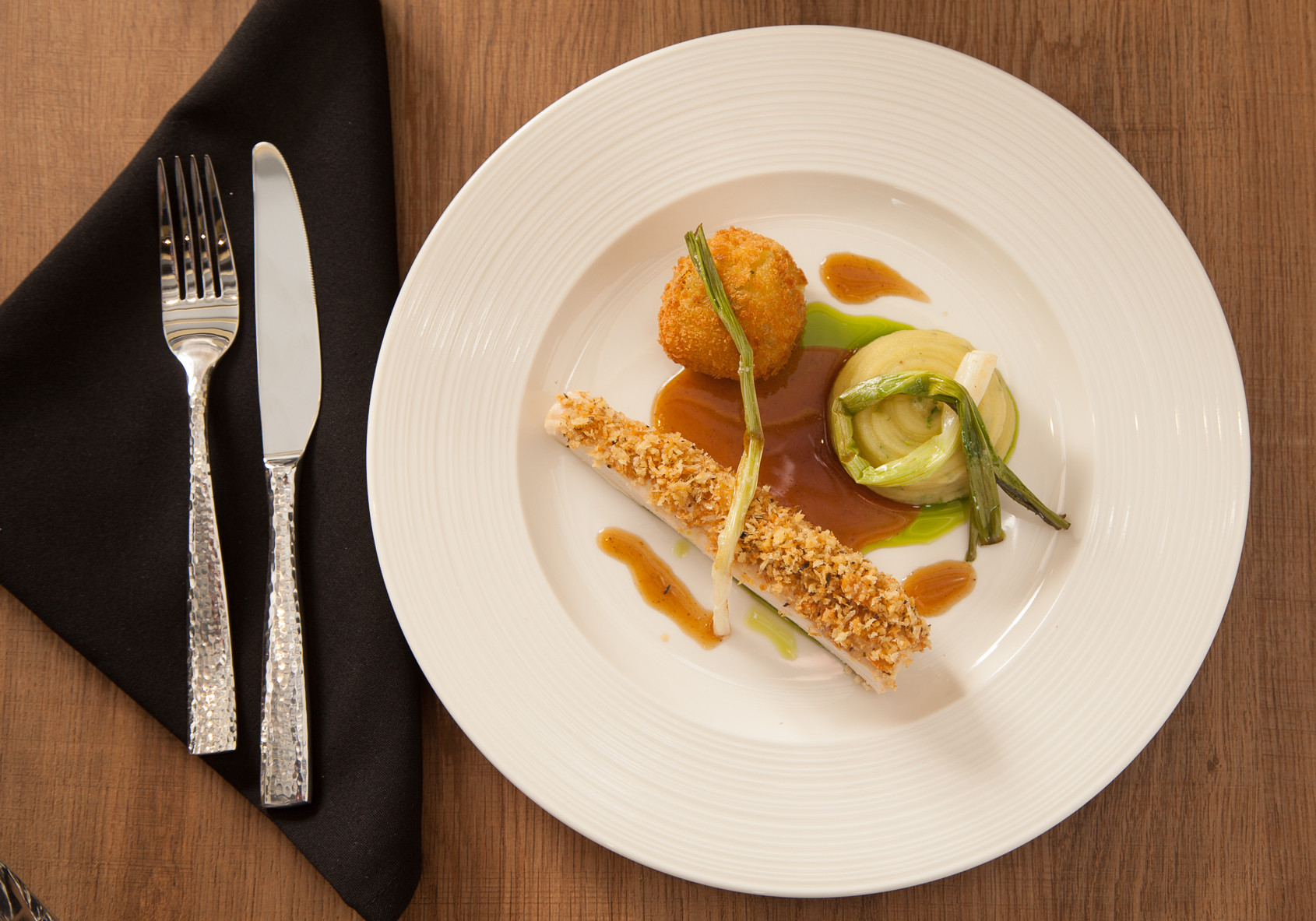 A elegantly plated dish featuring a crusted protein alongside a round fritter, garnished with green onions and a bed of creamy green puree. Decorative sauces and a drizzle of sauce complete the presentation. A fork and knife are placed beside the plate on a wooden table.