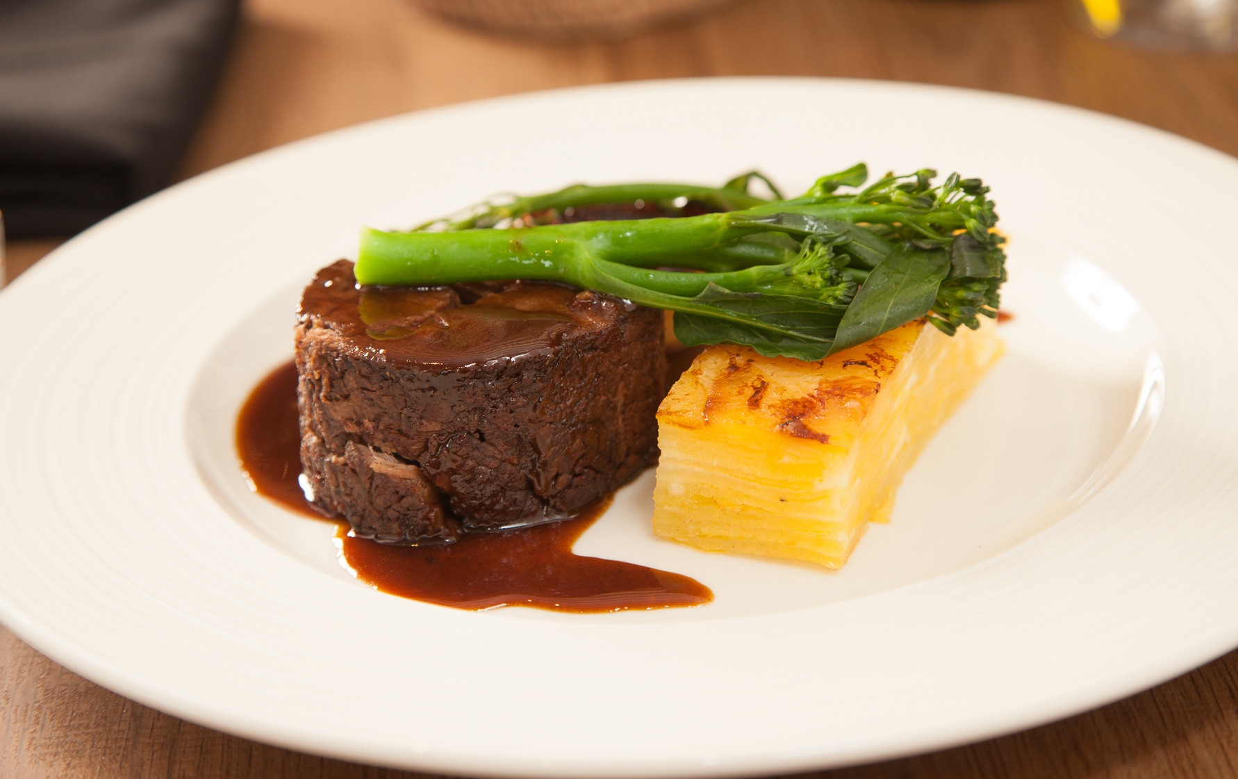 A gourmet dish featuring a round slice of tender beef, topped with rich brown sauce, accompanied by a portion of golden, layered potato and vibrant green broccoli stems.