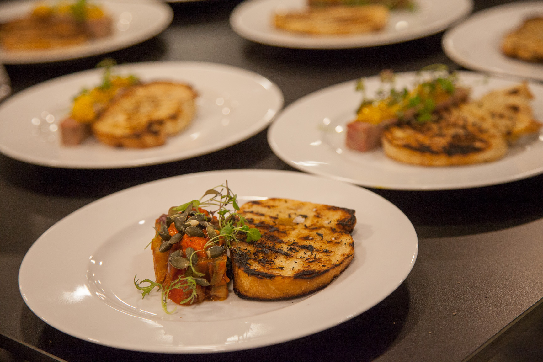 A white plate features a grilled slice of bread alongside a colorful vegetable medley topped with microgreens. Additional plates in the background display different grilled dishes.