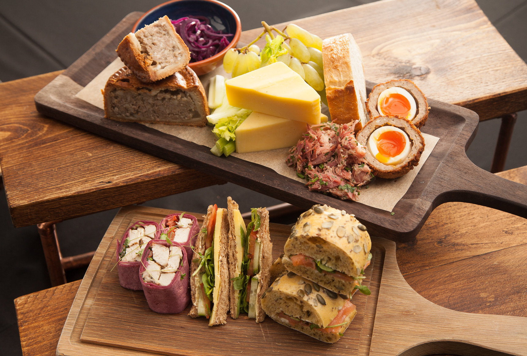 A wooden serving board displays a variety of foods including slices of cheese, pickled red cabbage, green grapes, a meat pie, scotch eggs, and a mixture of sandwiches with colorful fillings.