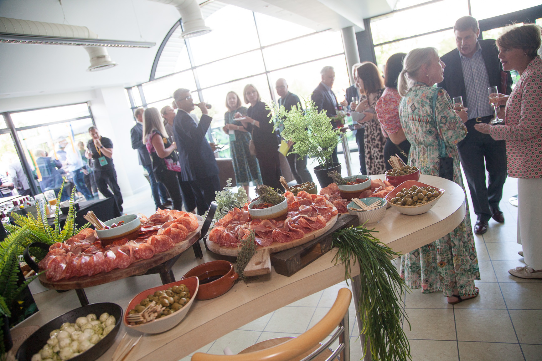 A well-decorated event space featuring guests mingling and enjoying refreshments. A display of charcuterie, including meats and olives, is set on a table in the foreground, complemented by greenery.