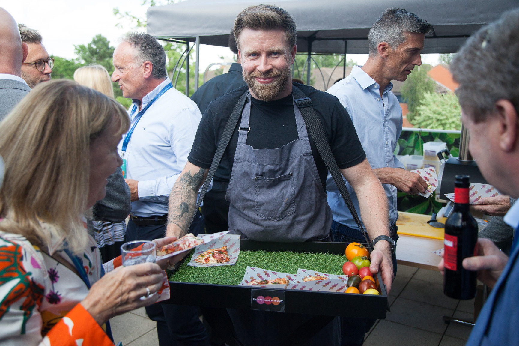 A man with a beard and tattoos wears an apron and smiles while holding a tray of food, including pizza slices and fruits, at an outdoor event with attendees mingling around him.