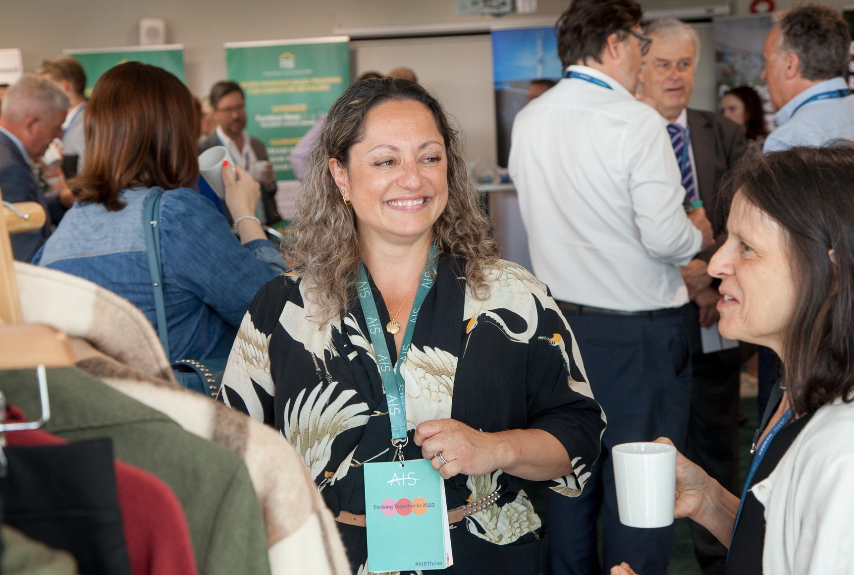 A woman with curly hair wearing a floral shirt smiles while holding a cup, engaging in conversation at a networking event. People are mingling in the background, with various banners and displays visible.