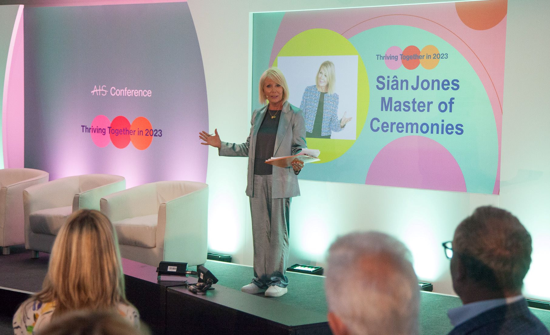 Sian Jones stands on stage as the Master of Ceremonies at the ATS Conference, engaging the audience with a smile. The backdrop features colorful graphics with the text 