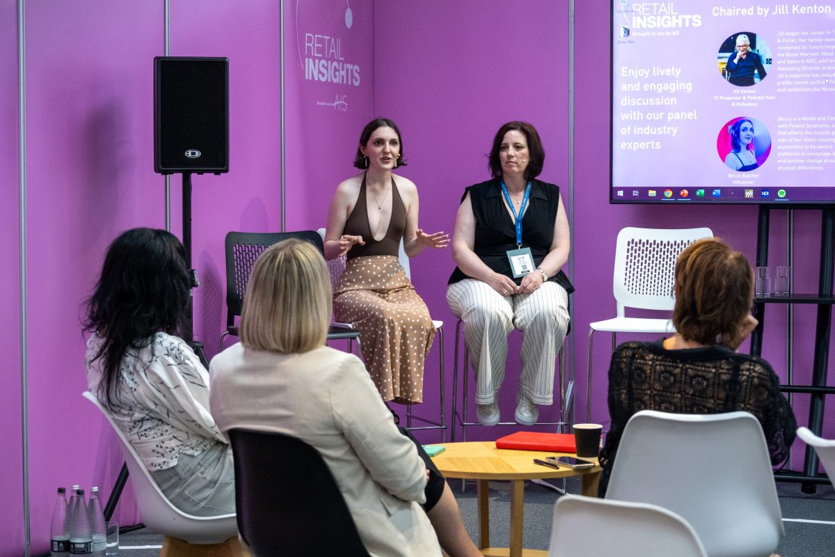 Two women are sitting on a stage, engaging in a discussion at a Retail Insights event. One woman is speaking animatedly while the other listens attentively. In the foreground, a small audience is seated, focused on the speakers. The backdrop features a vibrant pink wall with text related to the event.