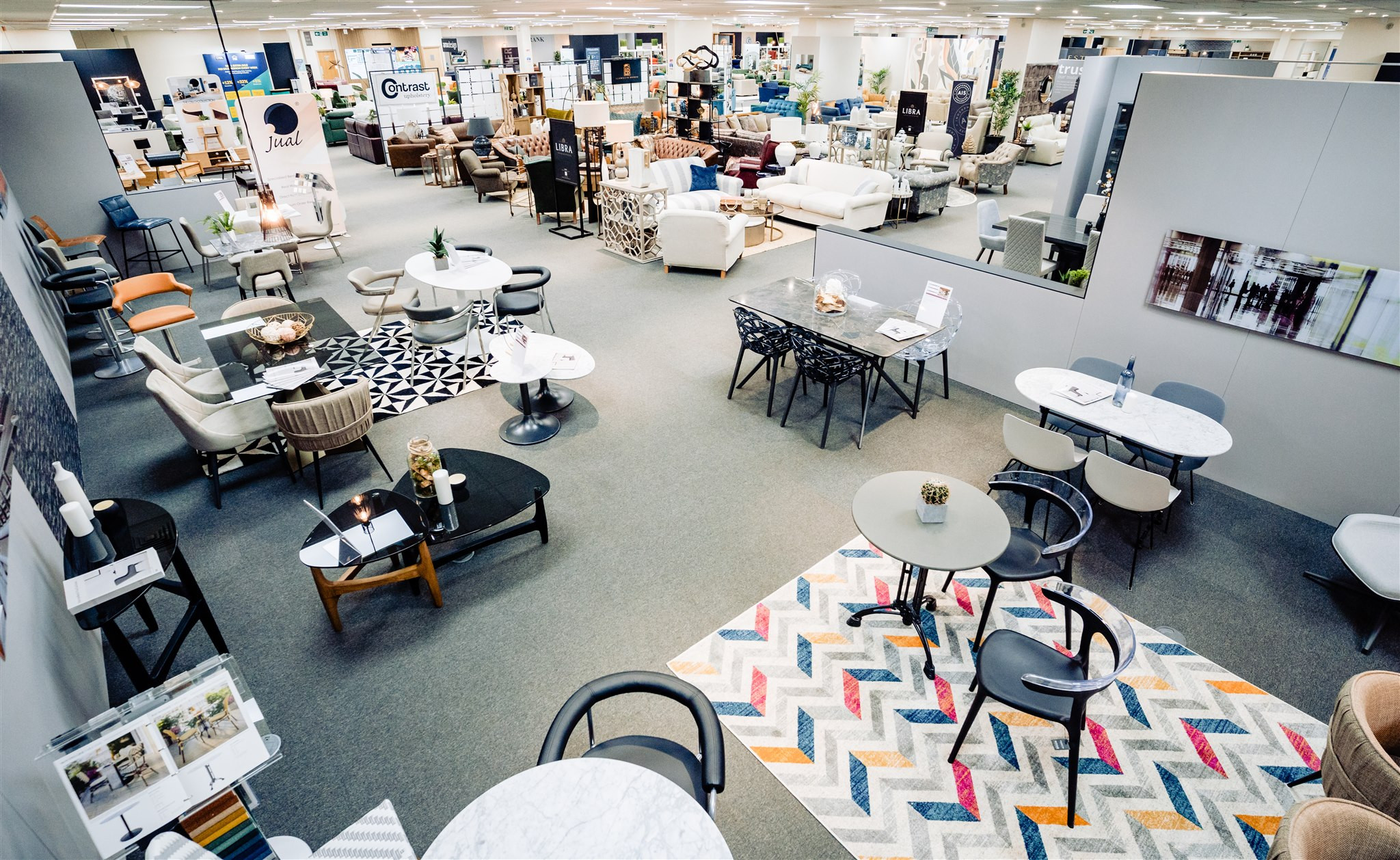 A modern furniture store interior showcases various seating arrangements and tables. Different styles of rugs add color to the space, while displays feature a mix of chairs and decorative items. The open layout highlights a range of furniture pieces for living and dining areas.