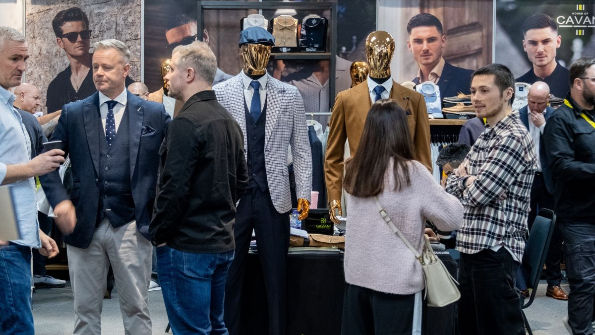 A crowd of people at a fashion exhibition, featuring well-dressed mannequins in stylish suits. Attendees are engaged in conversation, surrounded by fashion displays and promotional materials.
