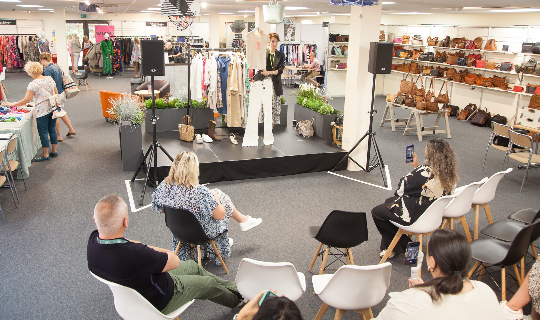 A fashion show is taking place indoors, featuring a model on a small stage wearing stylish clothing. The audience, seated on modern chairs, is attentively watching. Clothing racks and bags are visible in the background, creating a vibrant retail atmosphere.