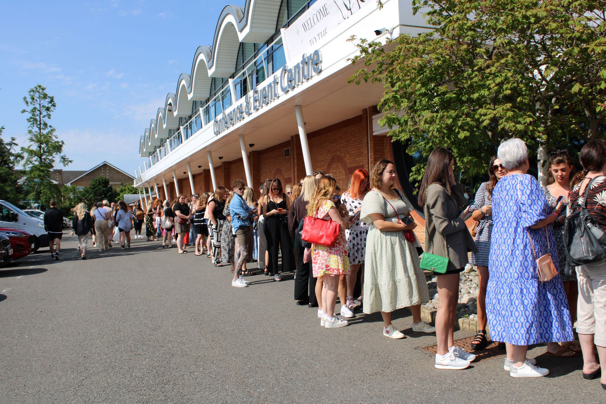 A long line of people waiting outside a building on a sunny day. The crowd is diverse, with individuals wearing various outfits, including dresses and casual attire. Trees and parked cars are visible along the pathway.