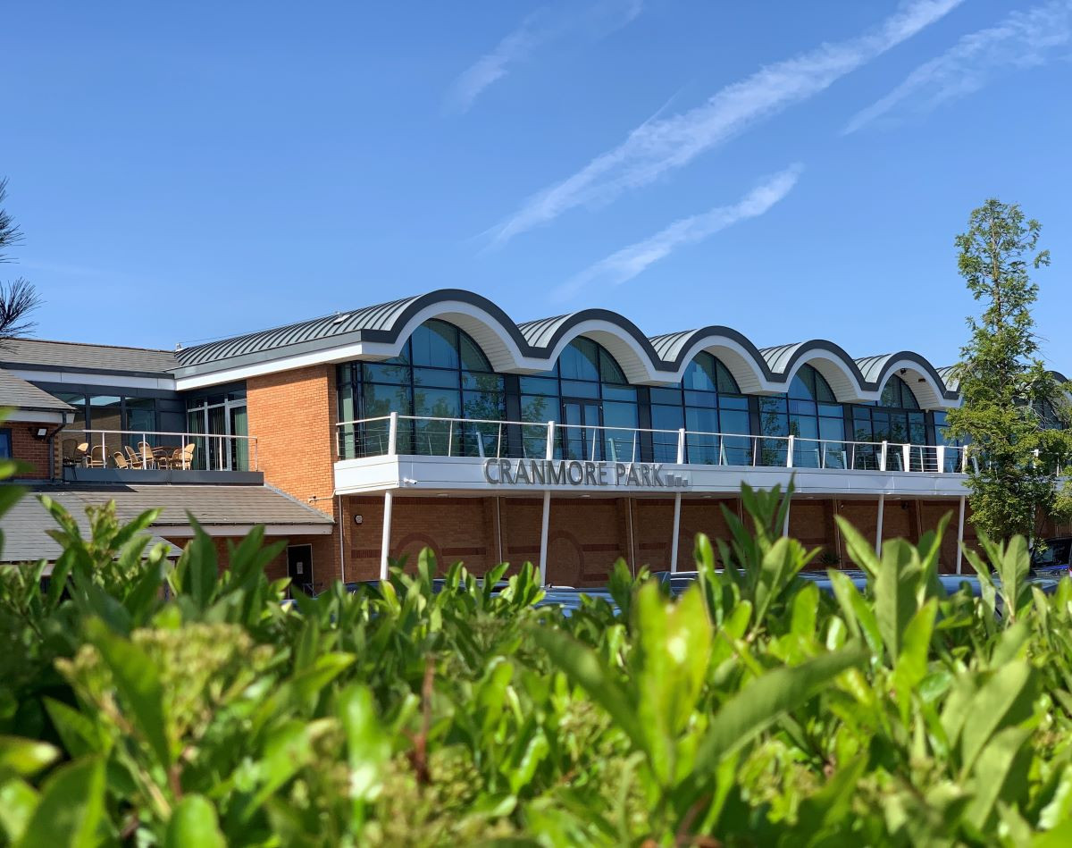 Cranmore Park building with a curved roof design, featuring large windows and a balcony. Clear blue sky and greenery in the foreground.