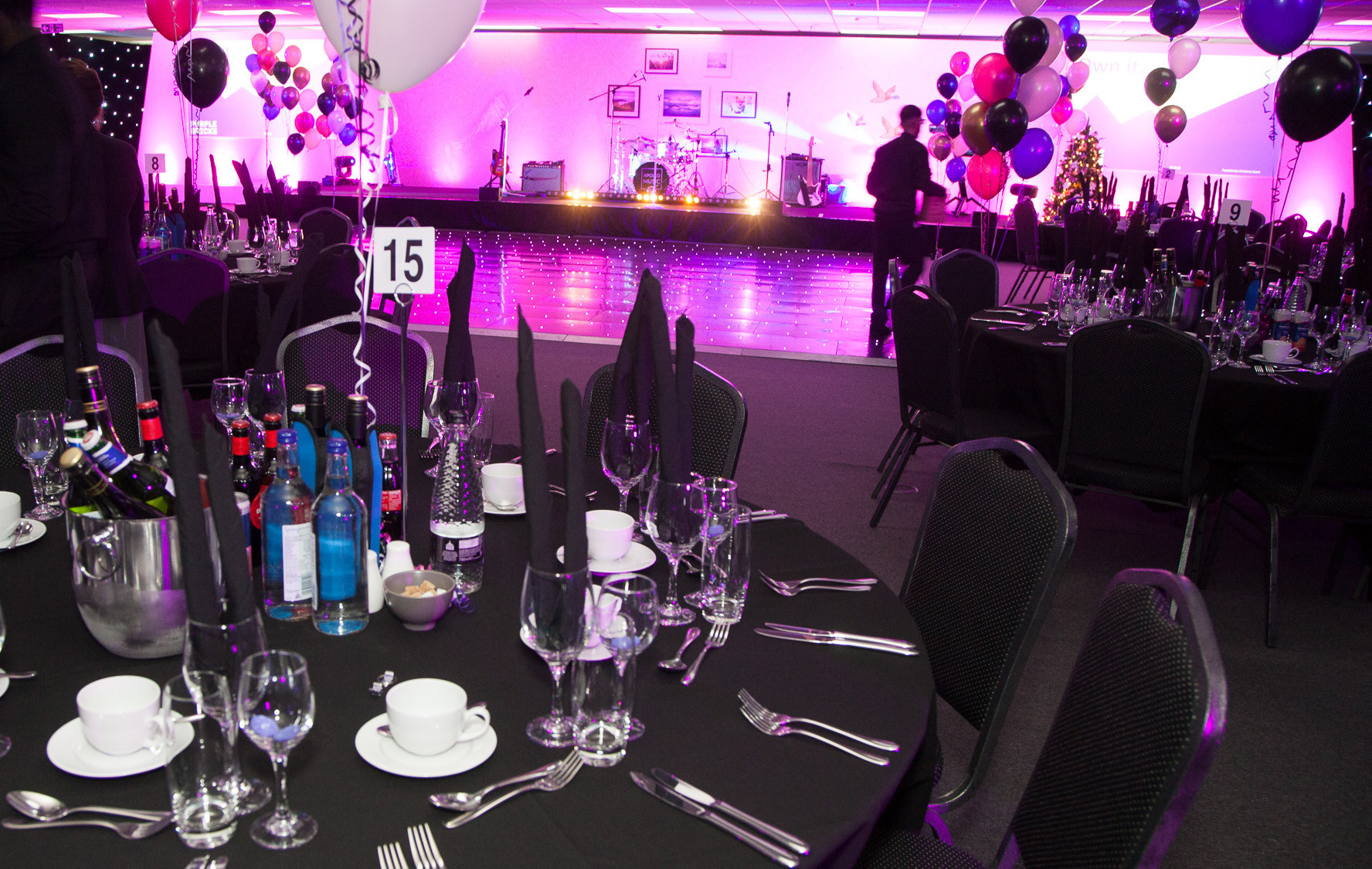 A formal dining setup featuring tables with black tablecloths, neatly arranged cutlery, and drinks. In the background, a stage with musical instruments is illuminated by colorful lights, and balloons in various colors are displayed around the room.