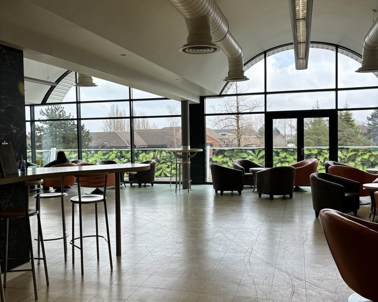 A spacious indoor seating area features modern furniture, including brown armchairs and high-top tables. Large windows provide natural light and a view of greenery outside. Soft, neutral-colored walls and a contemporary ceiling design create a welcoming atmosphere.