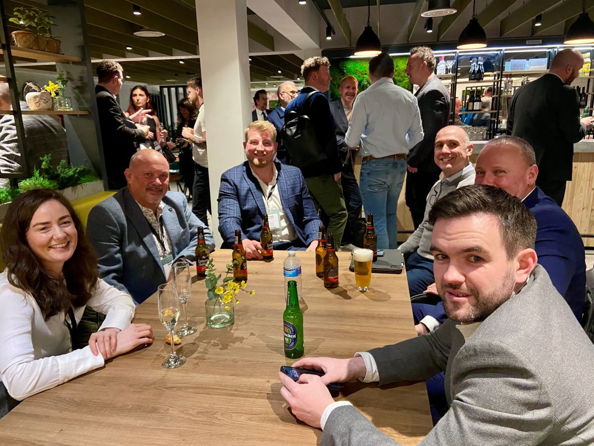 A group of six people sits around a table in a lively indoor setting. They are enjoying drinks, with visible beers and glasses. The background features other guests mingling and a bar area. Everyone is smiling and engaged in conversation.