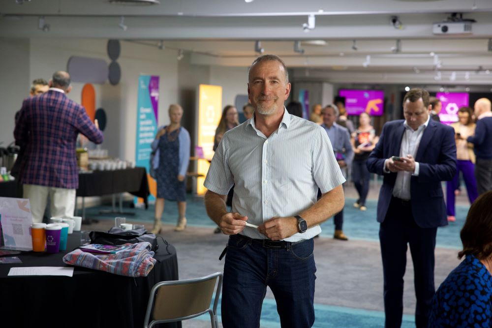 A man in a light blue shirt stands confidently in a modern event space, smiling at the camera. In the background, attendees engage in conversation and coffee service is visible. Bright banners decorate the room, adding to the lively atmosphere.