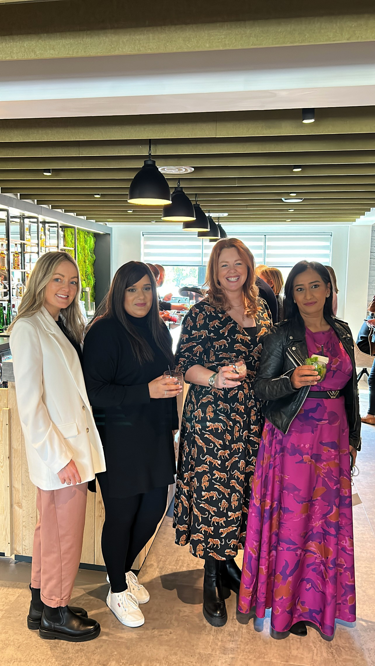 Four women stand together in a modern setting, each holding a drink. They are dressed in stylish outfits, with two in dresses and the others in a blazer and casual wear. The background features a bar area with greenery.