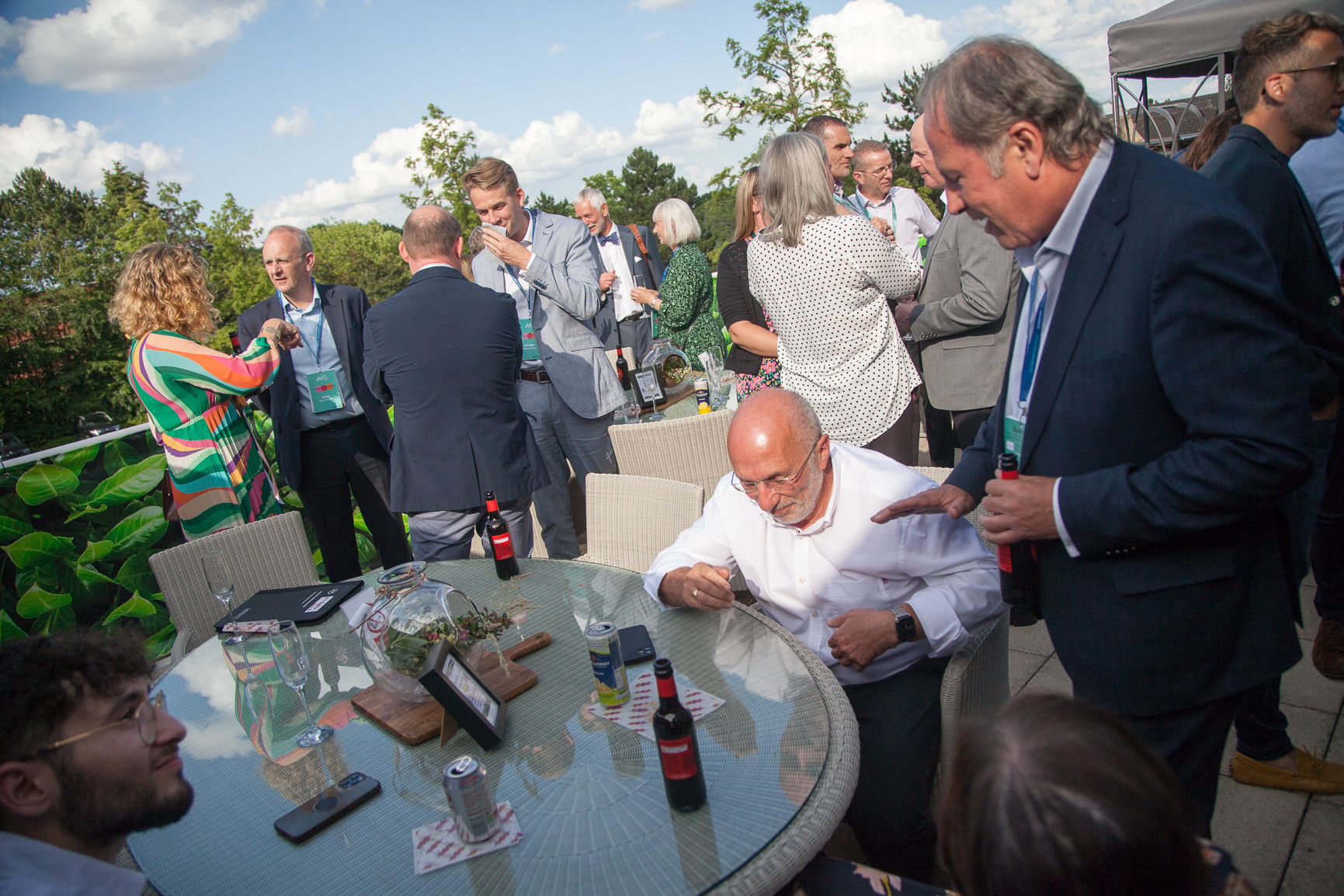A lively outdoor gathering with people mingling around a glass table. Some individuals are seated, while others stand in conversation, enjoying refreshments. The background features green trees and a partly cloudy sky, creating a relaxed atmosphere.