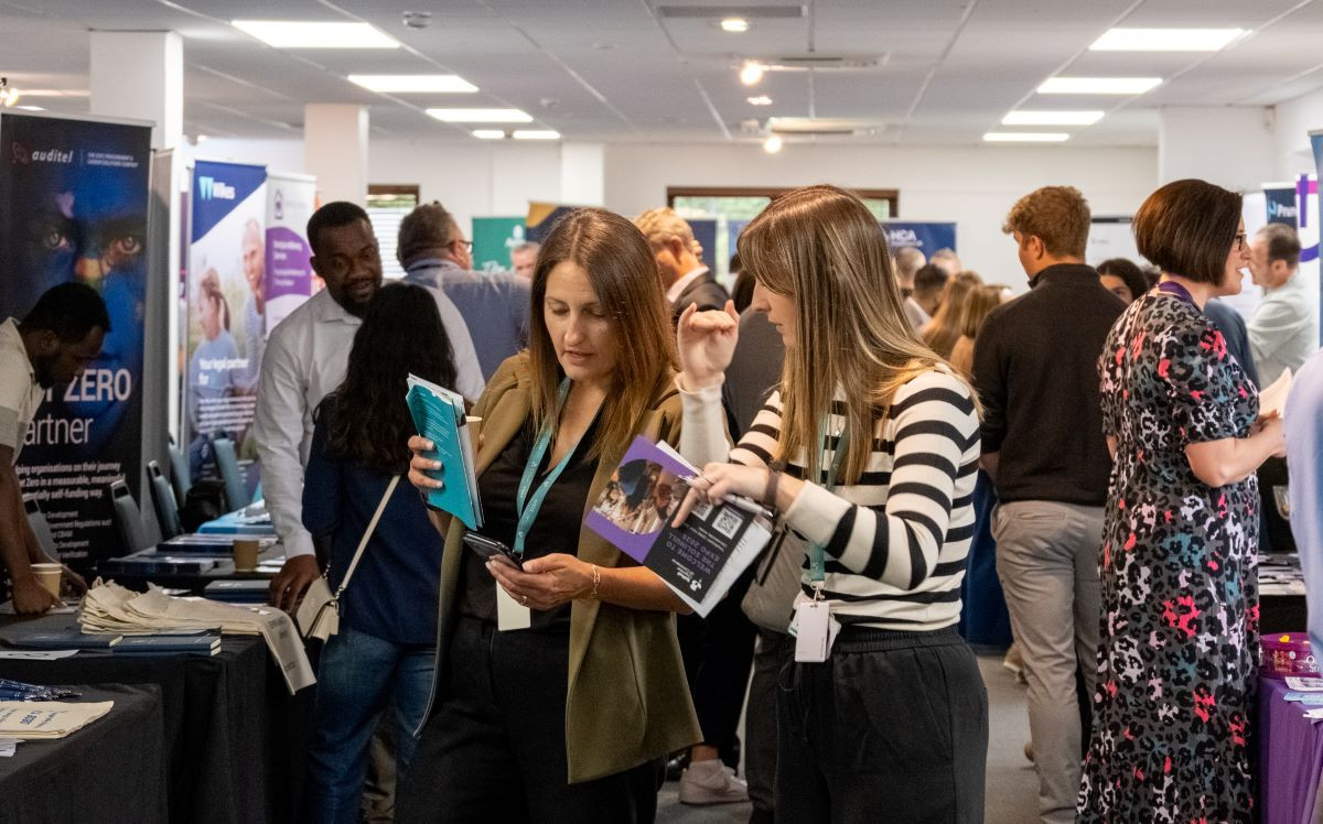 A busy event space with various vendors and attendees engaged in conversations. Two women in the foreground are examining brochures and materials, while people browse booths in the background. Promotional banners and tables are visible around the room.