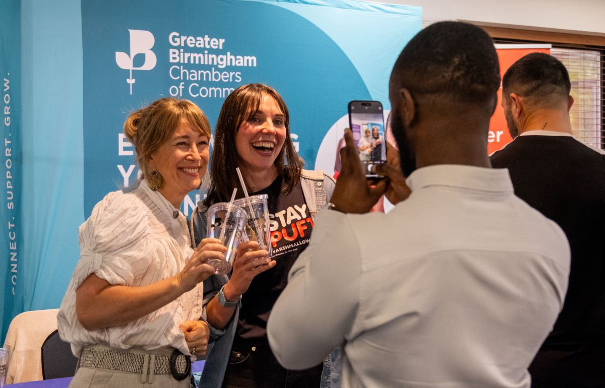 Two women smile and pose together, holding drinks, while a man takes their picture with a smartphone. They are standing in front of a backdrop that features the Greater Birmingham Chambers of Commerce logo.