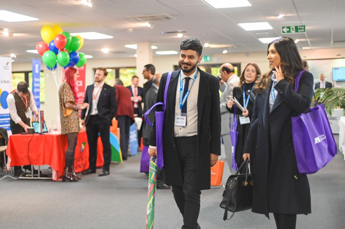 A group of professionals mingles at a networking event, with individuals carrying colorful bags. Balloons are in the background, and various exhibitor tables are set up around the venue.