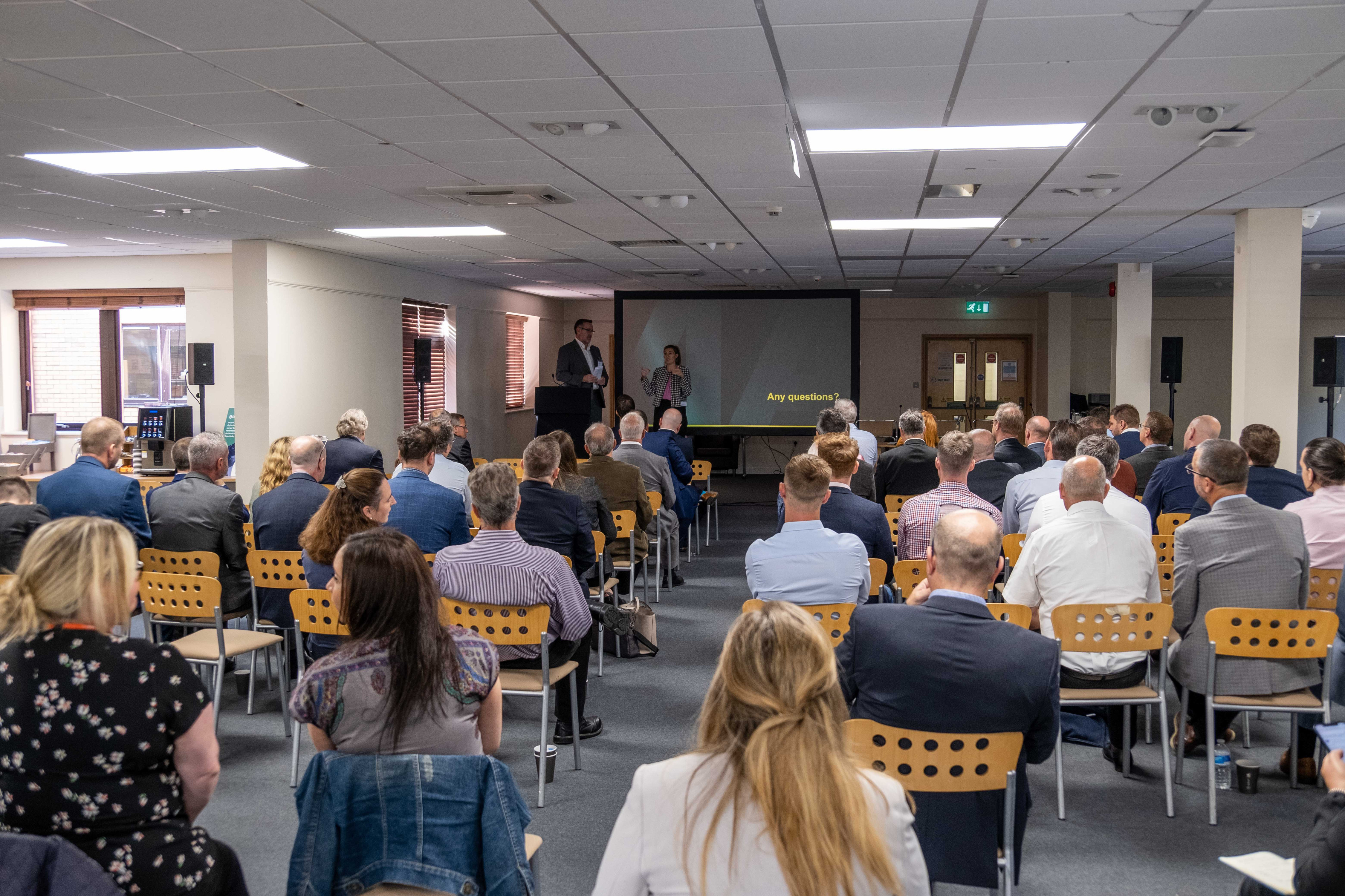 A crowded conference room with attendees seated, facing a presentation screen. Two speakers stand at the front, engaging the audience. Various people in business attire are positioned throughout the room, with a mix of men and women visible.
