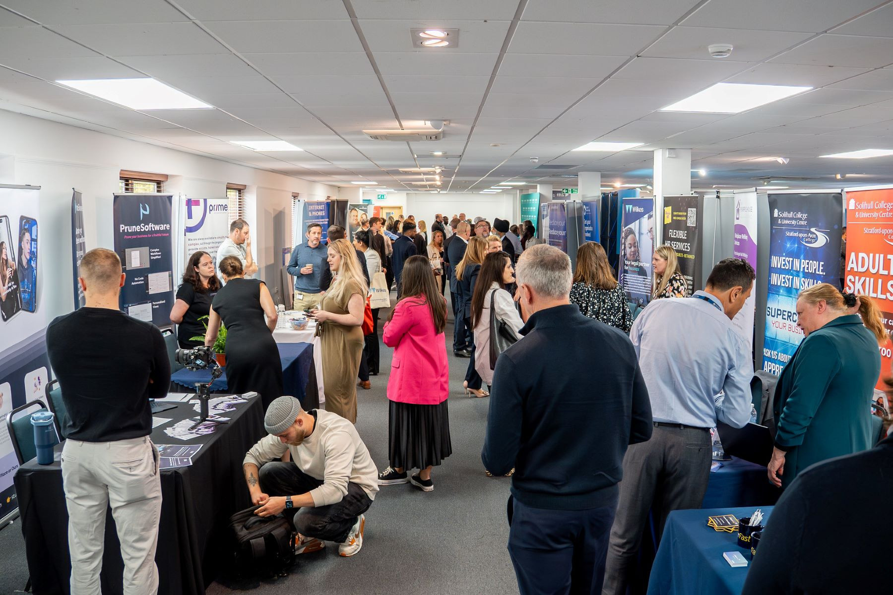 A bustling conference hall filled with people engaging at various booths. Attendees are networking and exploring displays about different programs and initiatives. Banners and signs promote services and skills training.