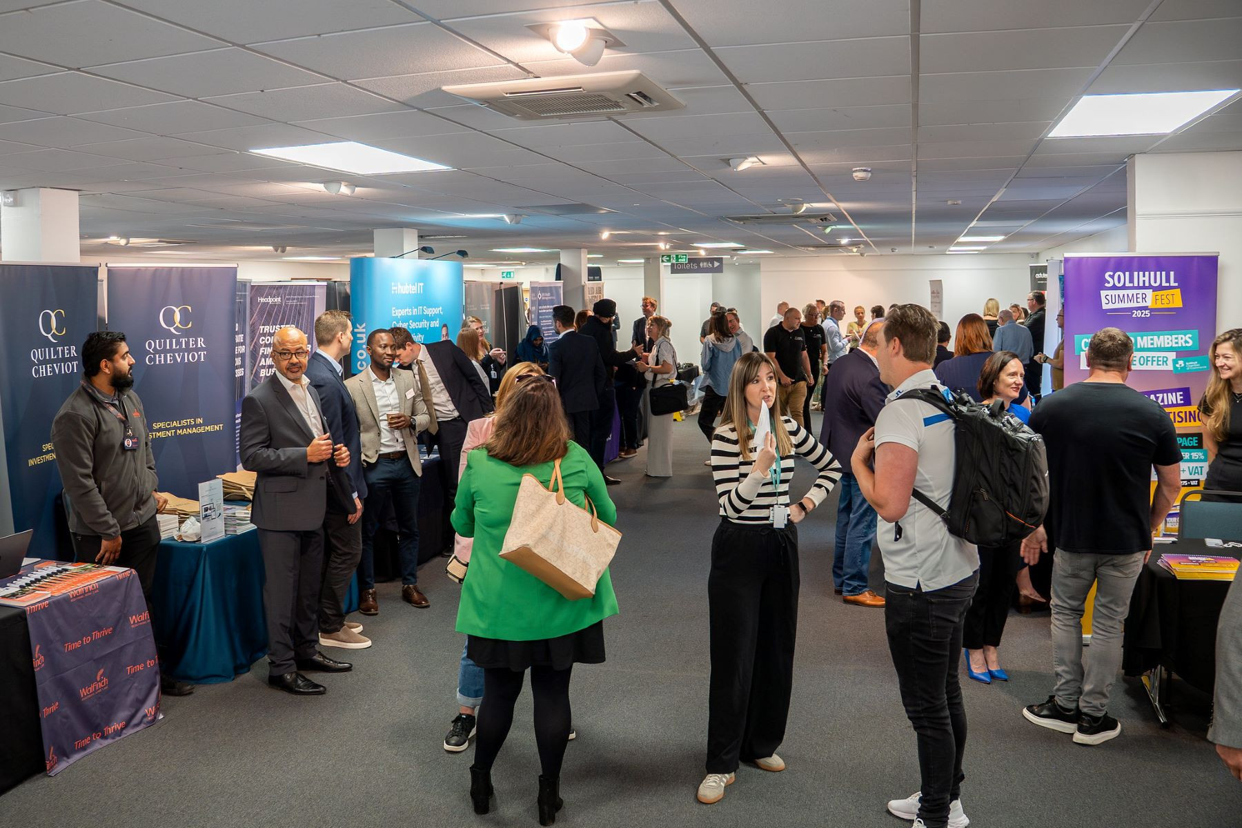 A bustling event space filled with people networking and engaging at various booths. Attendees are dressed in professional attire, with banners and tables showcasing different businesses and services.