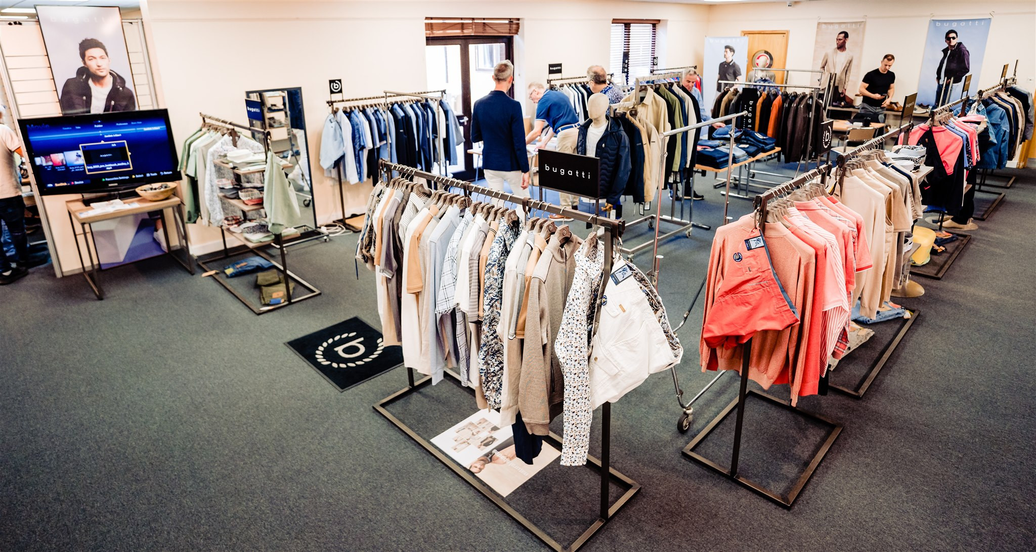A clothing showroom featuring racks of various shirts, jackets, and pants. Several customers browse the items, while a TV screen displays images of clothing. The space has neutral-colored walls and a carpeted floor.